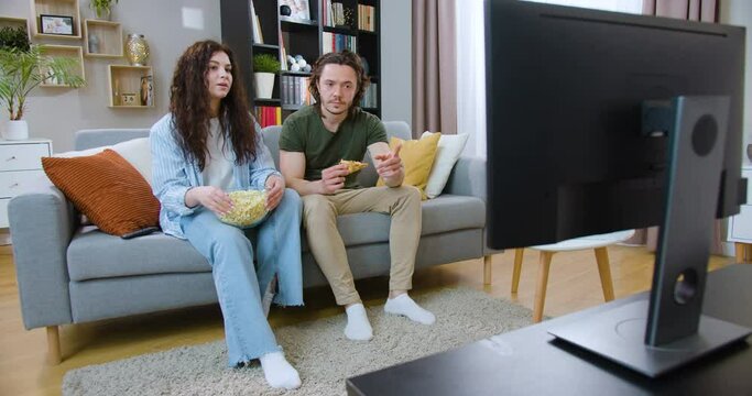 Attractive woman with curly dark hair switching channels on TV while sitting on couch with her boyfriend. Young couple eating pizza and popcorn and choosing entertainment program on television.