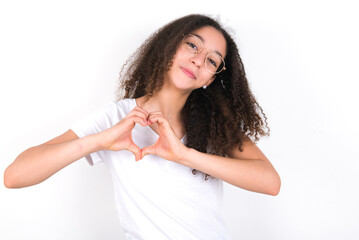 young beautiful girl with afro hairstyle wearing white t-shirt over white wall smiling in love showing heart symbol and shape with hands. Romantic concept.