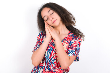 Relax and sleep time. Tired young beautiful girl with afro hairstyle wearing flowered dress with closed eyes leaning on palms making sleeping gesture.