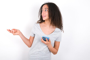 Happy pleased young beautiful girl with afro hairstyle wearing grey t-shirt over white wall raises palm and holds cellphone uses high speed internet for text messaging or video calls