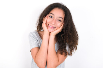 Happy young beautiful girl with afro hairstyle wearing grey t-shirt over white wall touches both cheeks gently, has tender smile, shows white teeth, gazes positively straightly at camera,