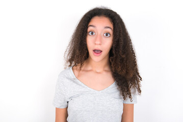 Shocked young beautiful girl with afro hairstyle wearing grey t-shirt over white wall stares bugged eyes keeps mouth opened has surprised expression. Omg concept