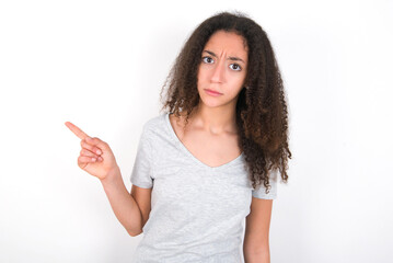 Serious young beautiful girl with afro hairstyle wearing grey t-shirt over white wall smirks face points away on copy space shows something unpleasant. Look at this advertisement. Big price concept