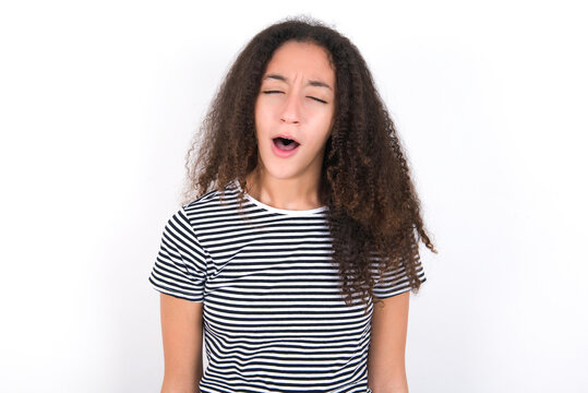 young beautiful girl with afro hairstyle wearing striped t-shirt over white wall yawns with opened mouth stands. Daily morning routine