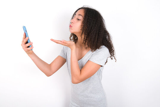 young beautiful girl with afro hairstyle wearing grey t-shirt over white wall  blows air kiss at camera of smartphone and takes selfie, sends mwah via online call.