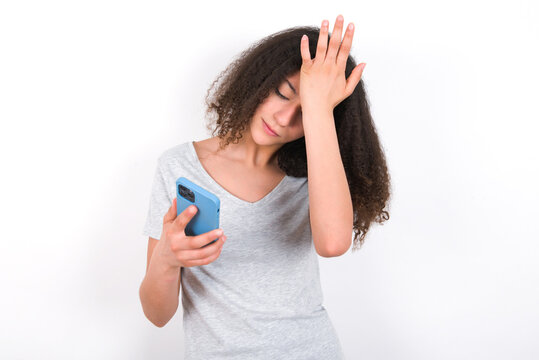 Upset depressed young beautiful girl with afro hairstyle wearing grey t-shirt over white wall makes face palm as forgot about something important holds mobile phone expresses sorrow and regret blames