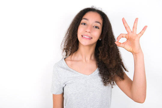 young beautiful girl with afro hairstyle wearing grey t-shirt over white wall hold hand arm okey symbol toothy approve advising novelty news