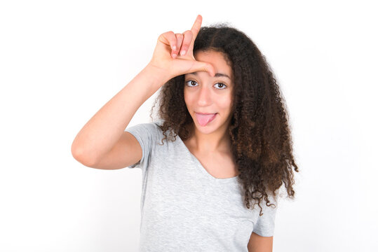 Funny young beautiful girl with afro hairstyle wearing grey t-shirt over white wall makes loser gesture mocking at someone sticks out tongue making grimace face.