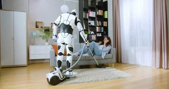 Futuristic cyborg using vacuum cleaner while woman in casual wear lying on comfy sofa and talking on smartphone at home. Concept of robot and human cooperation.