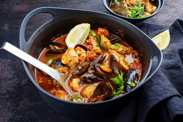 Modern style traditional Spanish seafood zarzuela de pescado with fish, king prawns and blue mussels served in red sauce as close-up in design pot