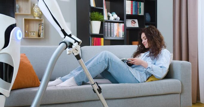Smiling young woman in casual wear lying on cozy couch and chatting with friends on wireless laptop during free time in modern apartment. Human like robot vacuuming floor at home.