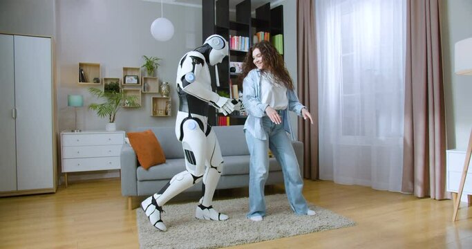 Full length portrait of attractive young woman dancing with bionic automated cyborg in living room. Humanoid robot and female with curly hair enjoying leisure activity indoors.