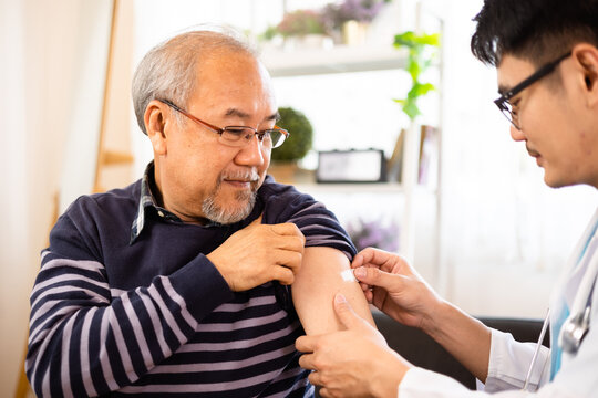 Safe vaccination for old people. Elder man in medical  getting flu or Covid-19 vaccine sitting on sofa at home. Asian Doctor or nurse giving flu or Covid-19 shot to senior.