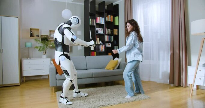 Carefree joyful woman dancing with human like futuristic robot in cozy apartment. Happy caucasian brunette in casual wear having fun and making high five gesture with cyborg.