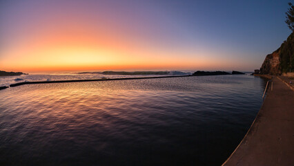 Obraz premium Beach dawn sunlight sky colors reflections over rocky tidal swimming pool with ocean sunrise nearby.