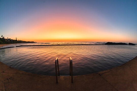 Beach dawn sunlight sky colors reflections over rocky tidal swimming pool with ocean sunrise nearby.