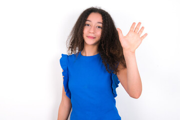 Teenager girl with afro hairstyle wearing blue T-shirt over white wall  Waiving saying hello happy and smiling, friendly welcome gesture.