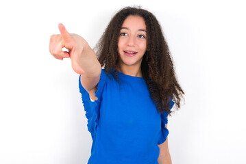 Teenager girl with afro hairstyle wearing blue T-shirt over white wall  Pointing with finger surprised ahead, open mouth amazed expression, something on the front.