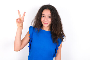 Fototapeta premium Teenager girl with afro hairstyle wearing blue T-shirt over white wall showing and pointing up with fingers number two while smiling confident and happy.
