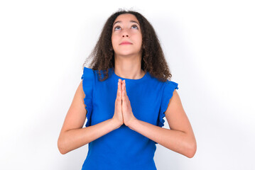 Teenager girl with afro hairstyle wearing blue T-shirt over white wall  begging and praying with...