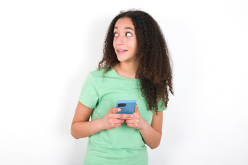 Teenager girl with afro hairstyle wearing green T-shirt over white wall holding a smartphone and looking sideways at blank copyspace.