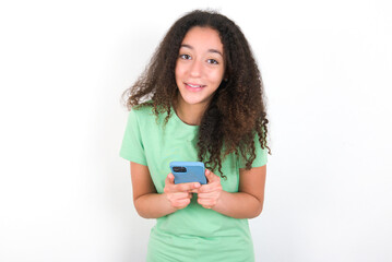 Excited Teenager girl with afro hairstyle wearing green T-shirt over white wall holding smartphone and looking amazed to the camera after receiving good news.