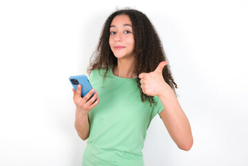 Portrait of Teenager girl with afro hairstyle wearing green T-shirt over white wall using and texting with smartphone  happy with big smile doing ok sign, thumb up with fingers, excellent sign