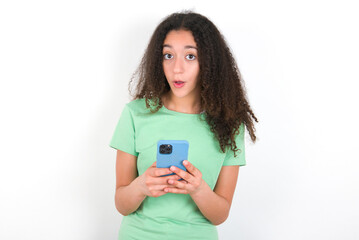 Shocked Teenager girl with afro hairstyle wearing green T-shirt over white wall opens mouth hold phone reading advert unbelievable big shopping prices