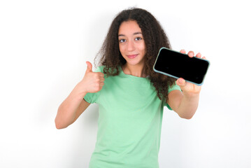 Portrait Teenager girl with afro hairstyle wearing green T-shirt over white wall holding in hands cell showing giving black screen thumb up