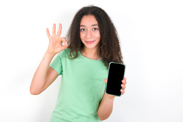 Teenager girl with afro hairstyle wearing green T-shirt over white wall holding in hands cell showing ok-sign