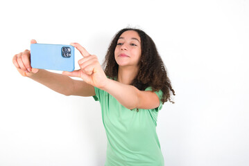 Teenager girl with afro hairstyle wearing green T-shirt over white wall taking a selfie to post it on social media or having a video call with friends.