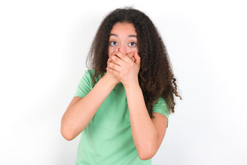Stunned Teenager girl with afro hairstyle wearing green T-shirt over white wall covers both hands on mouth, afraids of something astonishing