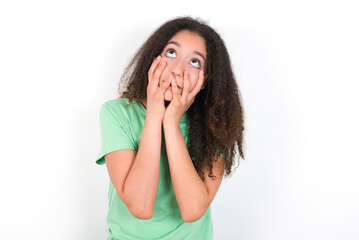 Teenager girl with afro hairstyle wearing green T-shirt over white wall keeps hands on cheeks has bored displeased expression. Stressed hopeless model