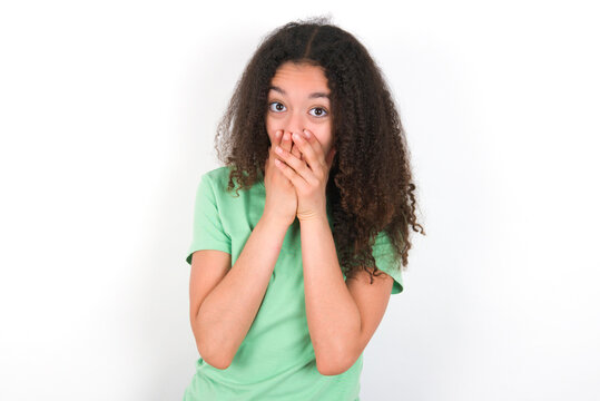 Teenager girl with afro hairstyle wearing green T-shirt over white wall keeps hands on mouth, looks with eyes full of disbelief, being puzzled with amount of work
