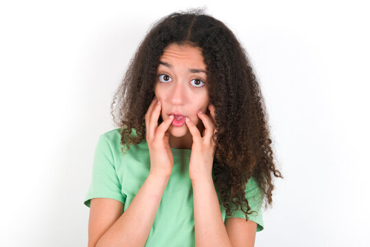Speechless Teenager girl with afro hairstyle wearing green T-shirt over white wall  keeps hands near opened mouth reacts to shocking news stares wondered at camera
