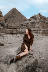 Young redhead tourist girl in brown dress sitting on a stone in Egypt, Cairo - Giza. Pyramids on backround. Copy space