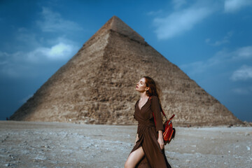 Young redhead tourist girl in brawn dress with a leather backpack standing on the sand in Egypt,...