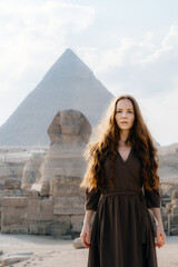 Young redhead woman standing in front of the Great Sphinx of Giza with the amazing Pyramid at the back. Valley of the Kings, Cairo, Egypt.