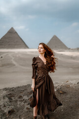 Young redhead tourist girl wearing a brown dress standing on the sand in Egypt, Cairo - Giza. Pyramids on backround. Copy space
