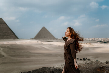 Young redhead tourist girl wearing a brown dress standing on the sand in Egypt, Cairo - Giza. Pyramids on backround. Copy space
