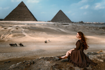 Young redhead tourist girl in brown dress sitting on a stone cliff in Egypt, Cairo - Giza. Pyramids on backround. Copy space
