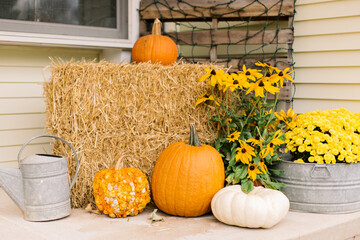 A decorative autumn front porch set up with pumpkins, gourds, mums, black eyed Susan’s, and a hay...