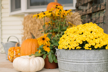 A decorative autumn front porch set up with pumpkins, gourds, mums, black eyed Susan’s, and a hay...