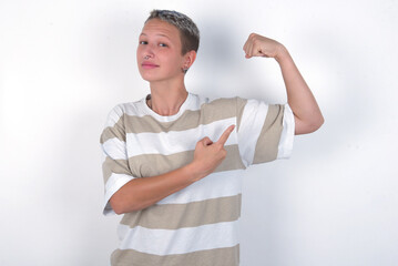 Smiling young woman with short hair wearing striped t-shirt over white background raises hand to show muscles, feels confident in victory, strong and independent.