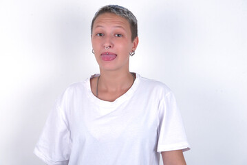 young woman with short hair wearing white t-shirt over white background with happy and funny face smiling and showing tongue.