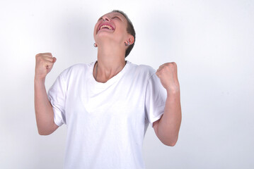 young woman with short hair wearing white t-shirt over white background looks with excitement up, keeps hands raised, notices something unexpected.