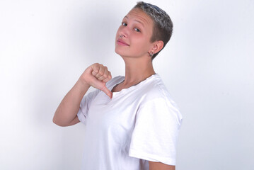 Closeup of cheerful young woman with short hair wearing white t-shirt over white background looks joyful, satisfied and confident, points at himself with thumb.