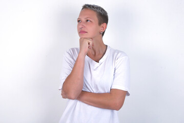 young woman with short hair wearing white t-shirt over white background with hand under chin and looking sideways with doubtful and skeptical expression, suspect and doubt.
