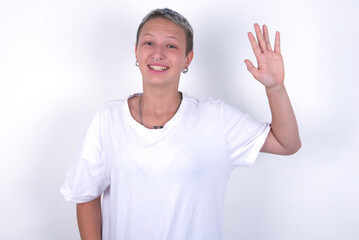 young woman with short hair wearing white t-shirt over white background waiving saying hello or...