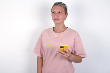 young woman with short hair wearing pink t-shirt over white background holds mobile phone uses high speed internet and social networks has online communication. Modern technologies concept